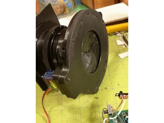 A black 3D-printed housing containing a leaf-shutter iris mechanism mounted on a camera lens, actuated by a small servo, on a cutting mat.